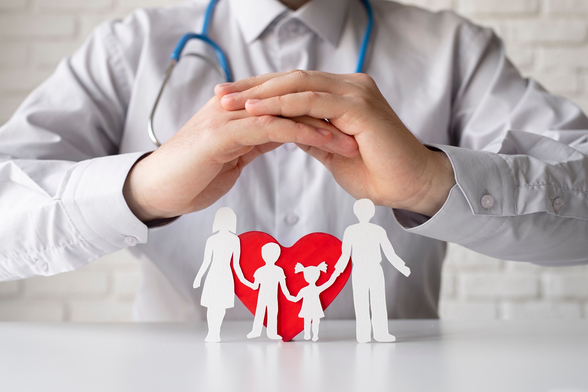 Stetoskoplu bir doktor, beyaz bir masa üzerindeki kırmızı bir kalbin önünde duran el ele tutuşmuş kağıttan aile figürlerinin üzerinde ellerini koruyucu bir şekilde birleştirmiş.